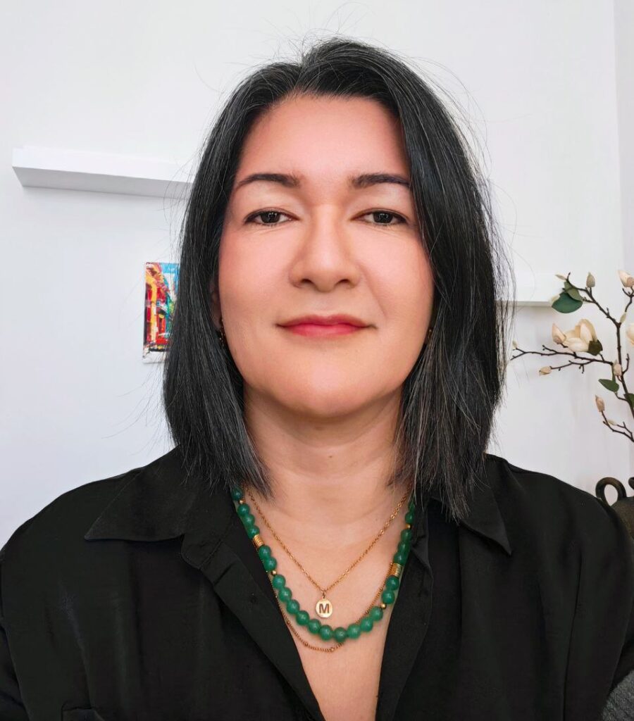 Una mujer con el pelo liso y oscuro hasta los hombros, con una camisa negra, un collar de cuentas verdes y un colgante dorado, está sentada en una habitación luminosa con una pared blanca y una decoración minimalista: un espacio inspirador para fortalecer tu confianza.